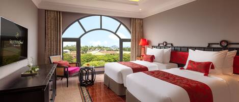 A hotel room with two beds, a TV, a desk, and a large window offering a view of the landscape.