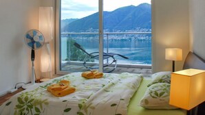 Habitación de hotel con cama, ventilador, lámpara y vistas a un lago y montañas.