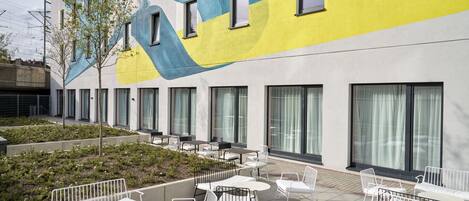 An outdoor seating area with white chairs and tables, a mural on the building, and a small garden.