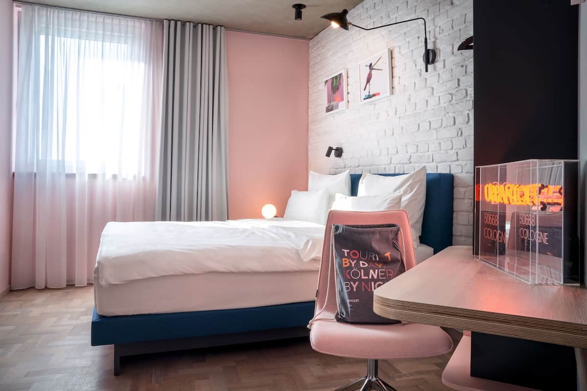 A modern hotel room with a bed, a desk, and a chair. There are curtains on the window, a wall clock, and a neon sign.