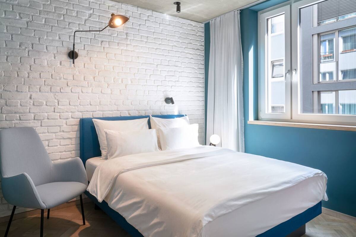 A room with a brick wall, a bed with white bedding, a blue headboard, a grey armchair, and a window with curtains.