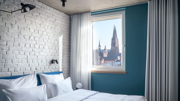 Habitación de hotel con pared de ladrillo blanco, un ventanal con vistas a una catedral y una cama con ropa blanca.