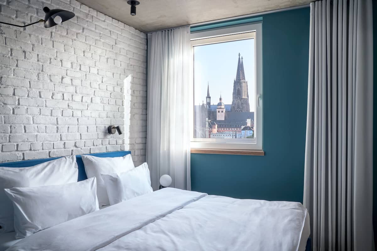 A hotel room with a white brick wall, a large window with a view of a cathedral, and a bed with white linens.
