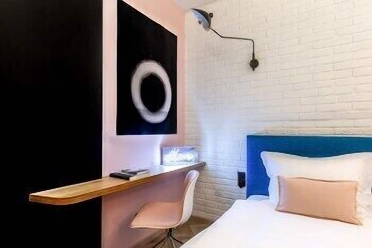 A modern hotel room with a bed, a desk, a chair, and a wall-mounted light fixture.