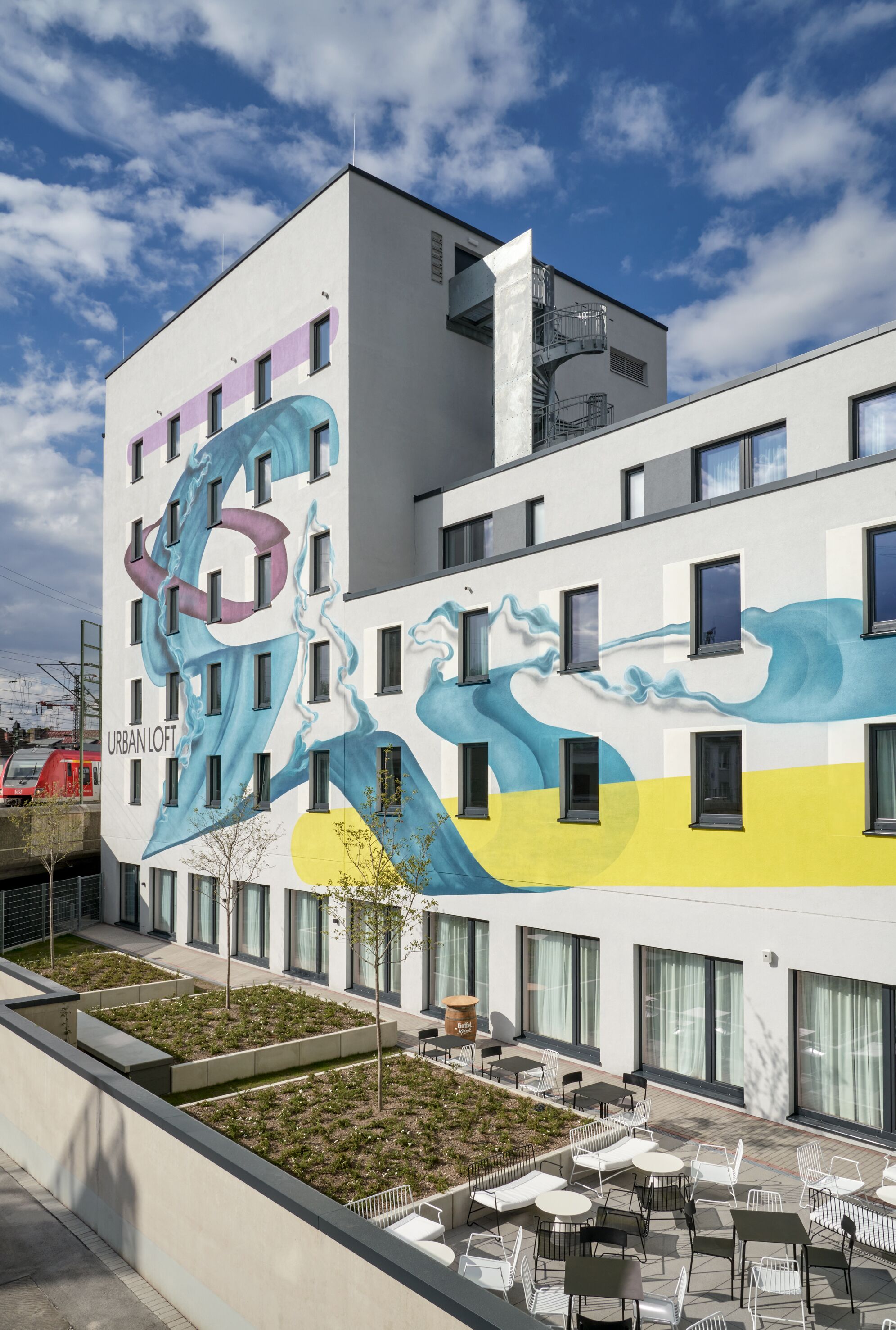 A modern building with a mural of a wave design on the facade, outdoor seating area, and a visible train in the background.