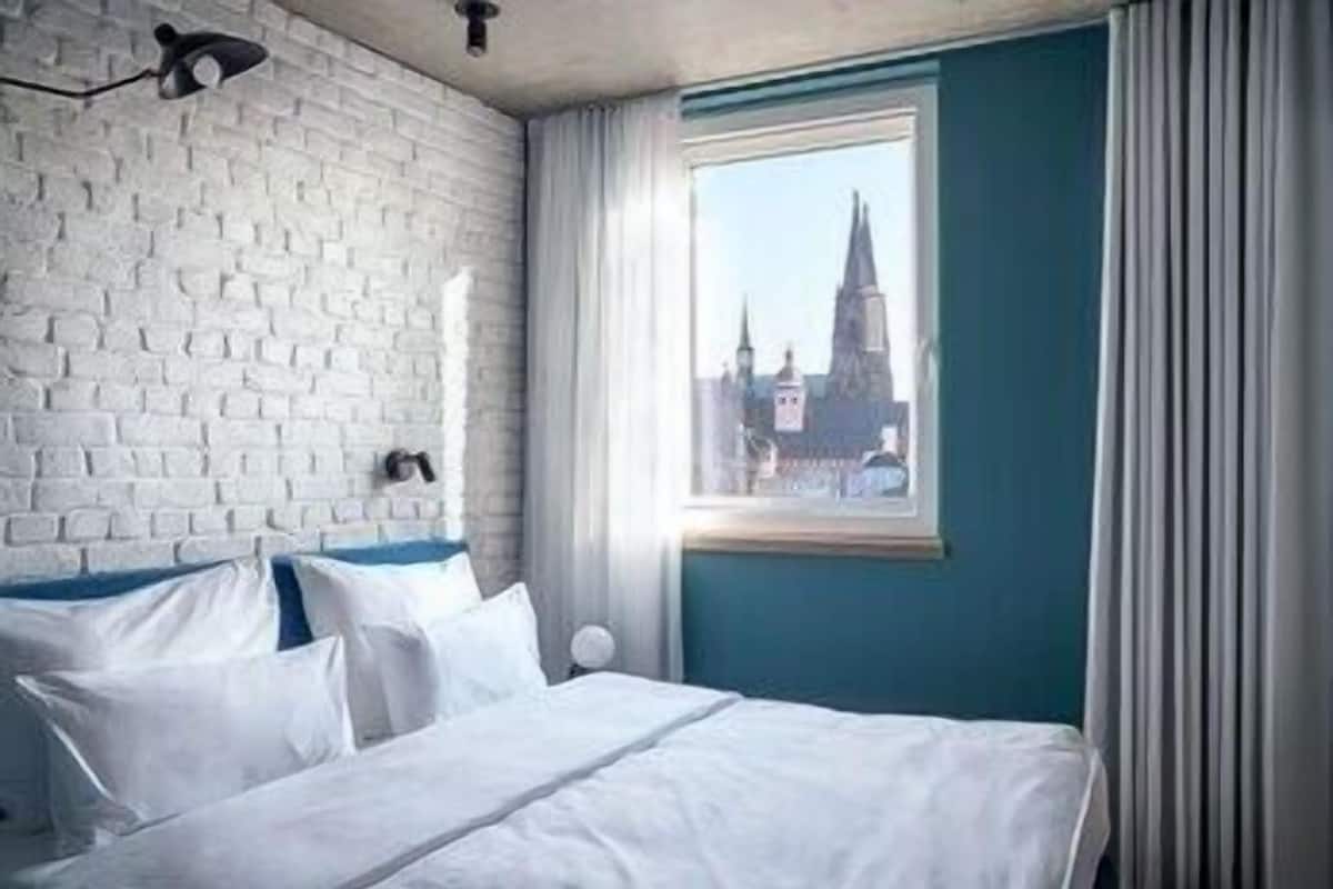 A hotel room with a white brick wall, a bed with white bedding, a window with a view of a cityscape, and a black wall-mounted lamp.