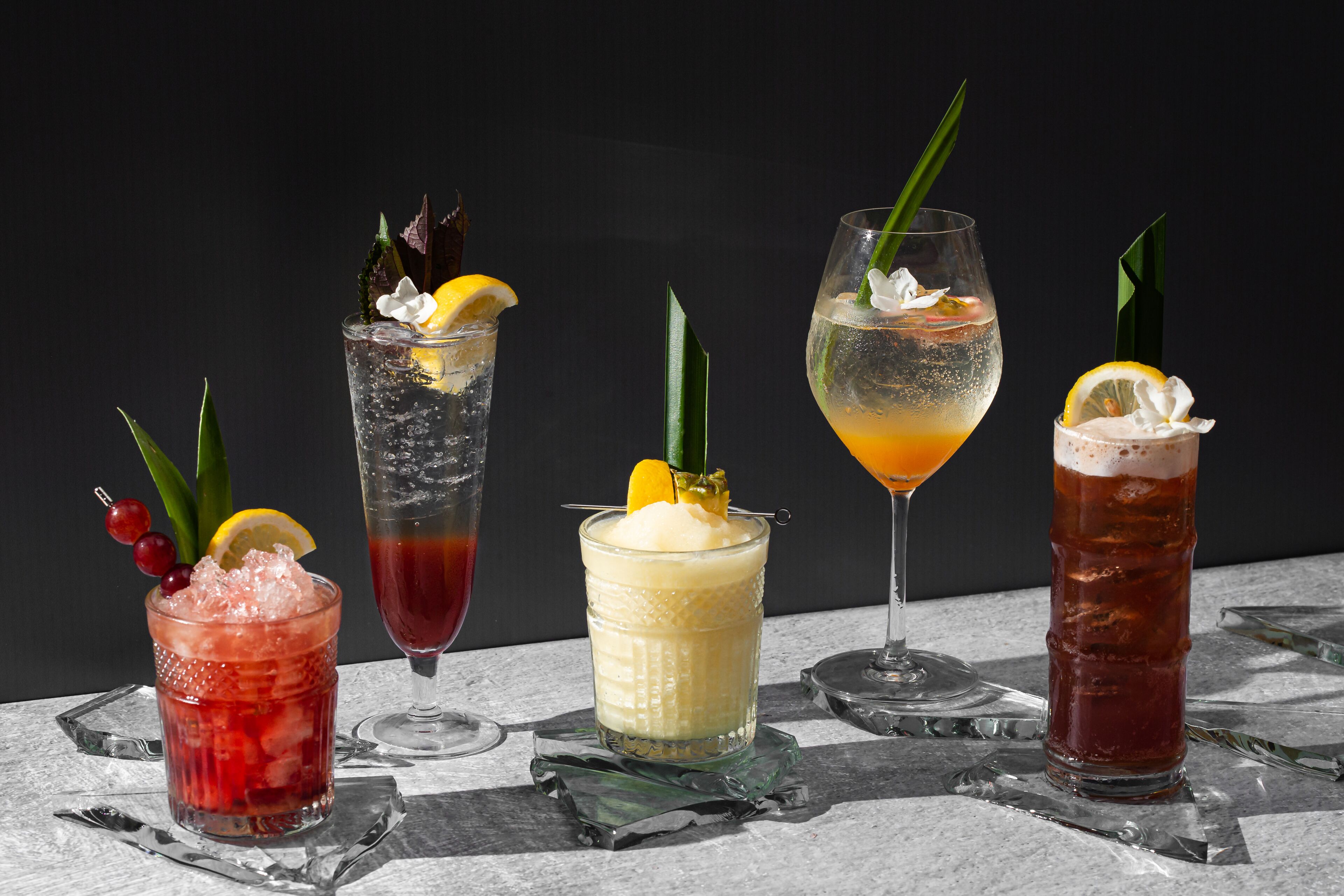 Five different cocktails with various garnishes and ice, displayed on a marble surface against a dark background.