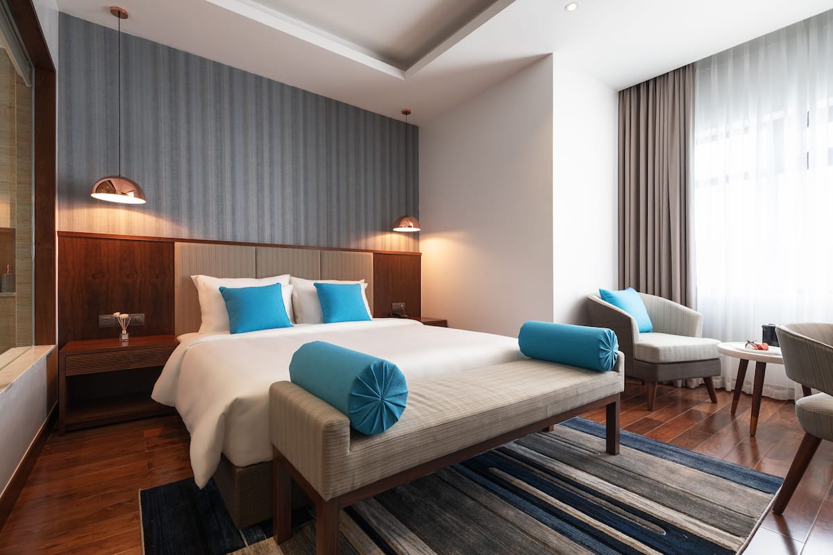 A modern hotel room with a large bed, a sitting area with a bench, a chair, and a small table. There are two bedside tables with lamps.