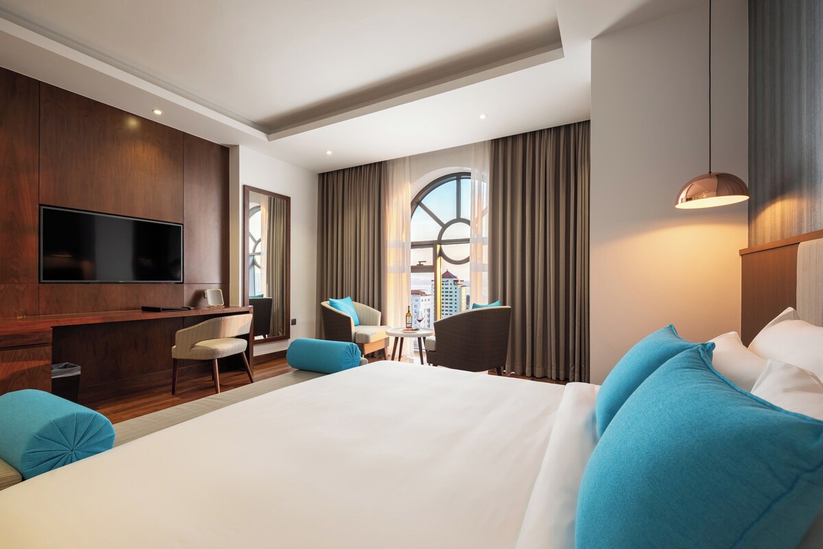 A hotel room with a large bed, a desk, a chair, a television, and a view of the city.