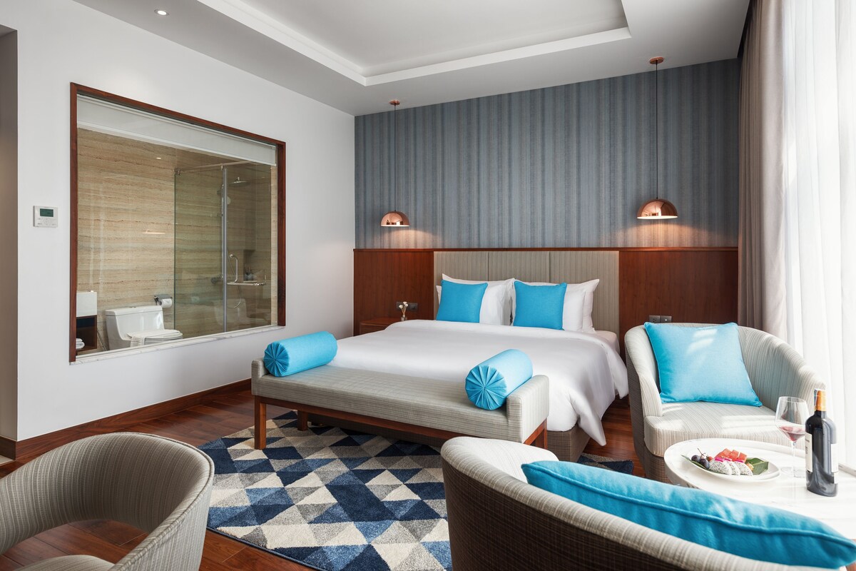 Habitación de hotel con una cama grande, dos mesitas de noche, una zona de estar con una silla y un sofá, cabina de ducha de vidrio y vistas al baño.
