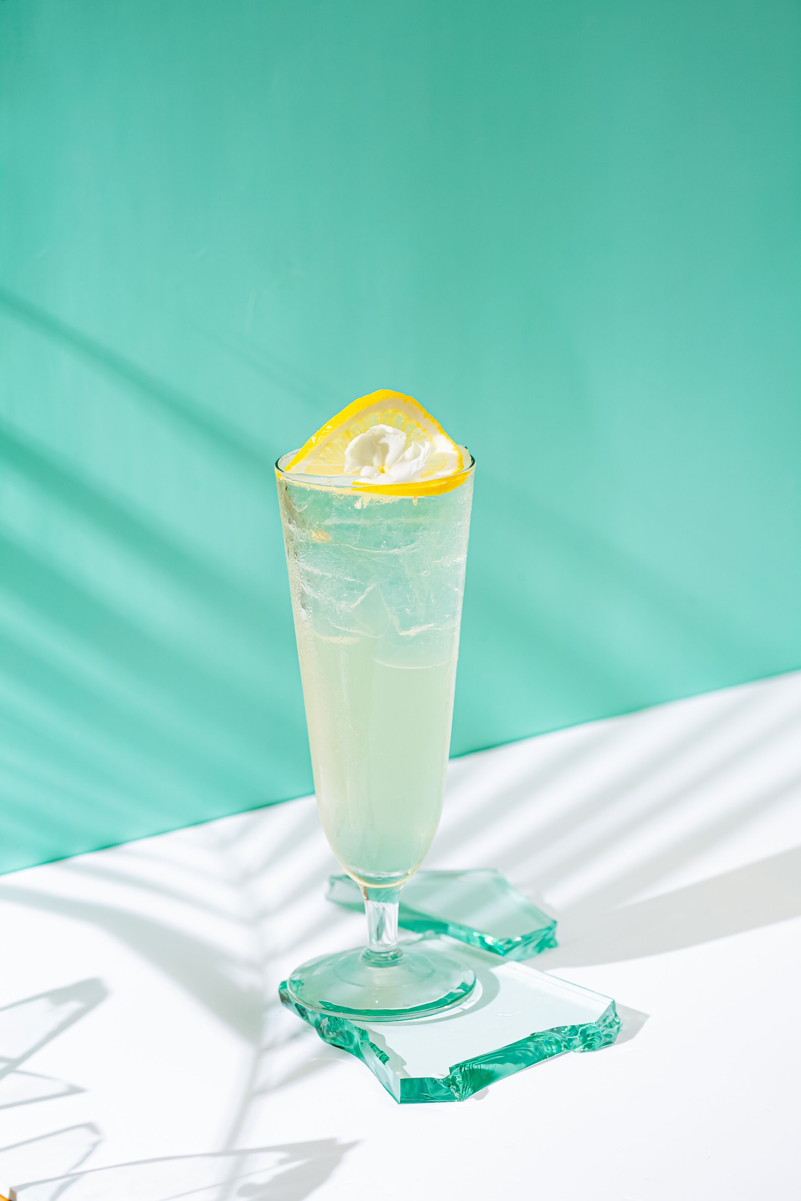A tall glass with a lemon slice garnish on a clear coaster, set against a teal background.
