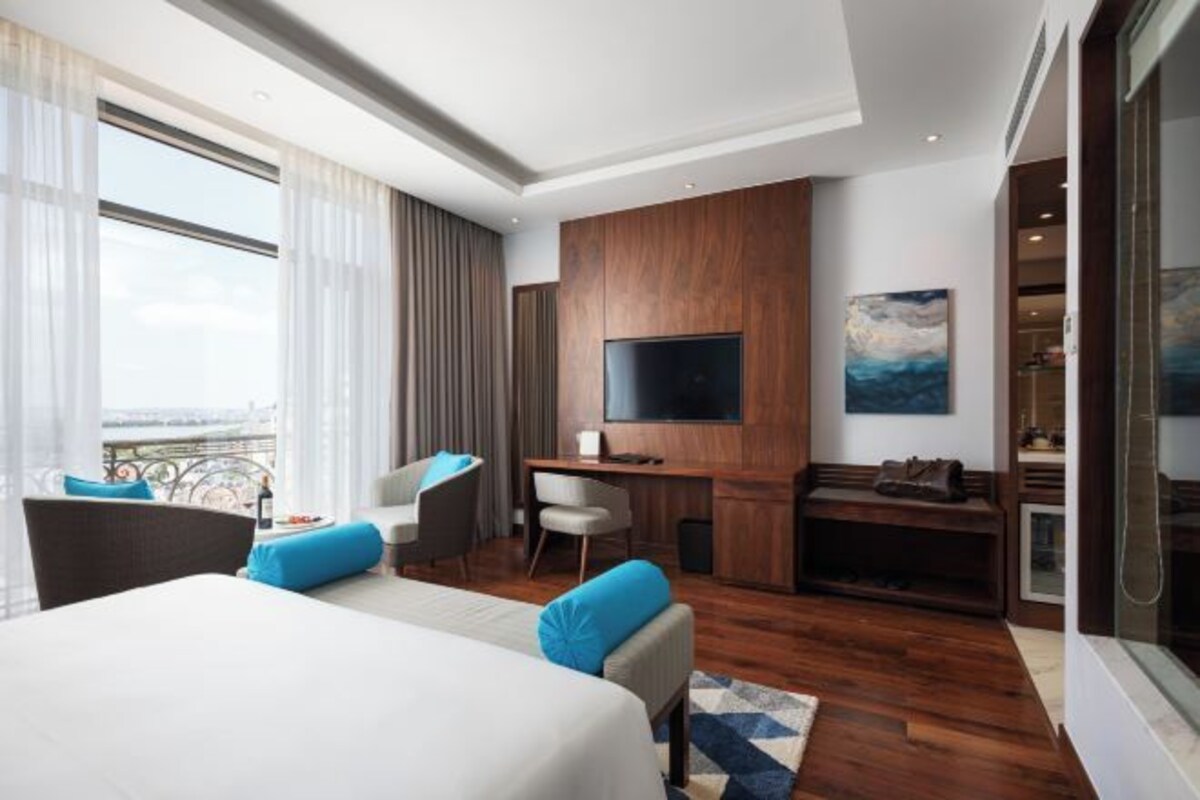 Habitación de hotel con una cama grande, un escritorio con televisor de pantalla plana, una zona de estar con sillas y vistas amplias a través de ventanas grandes.