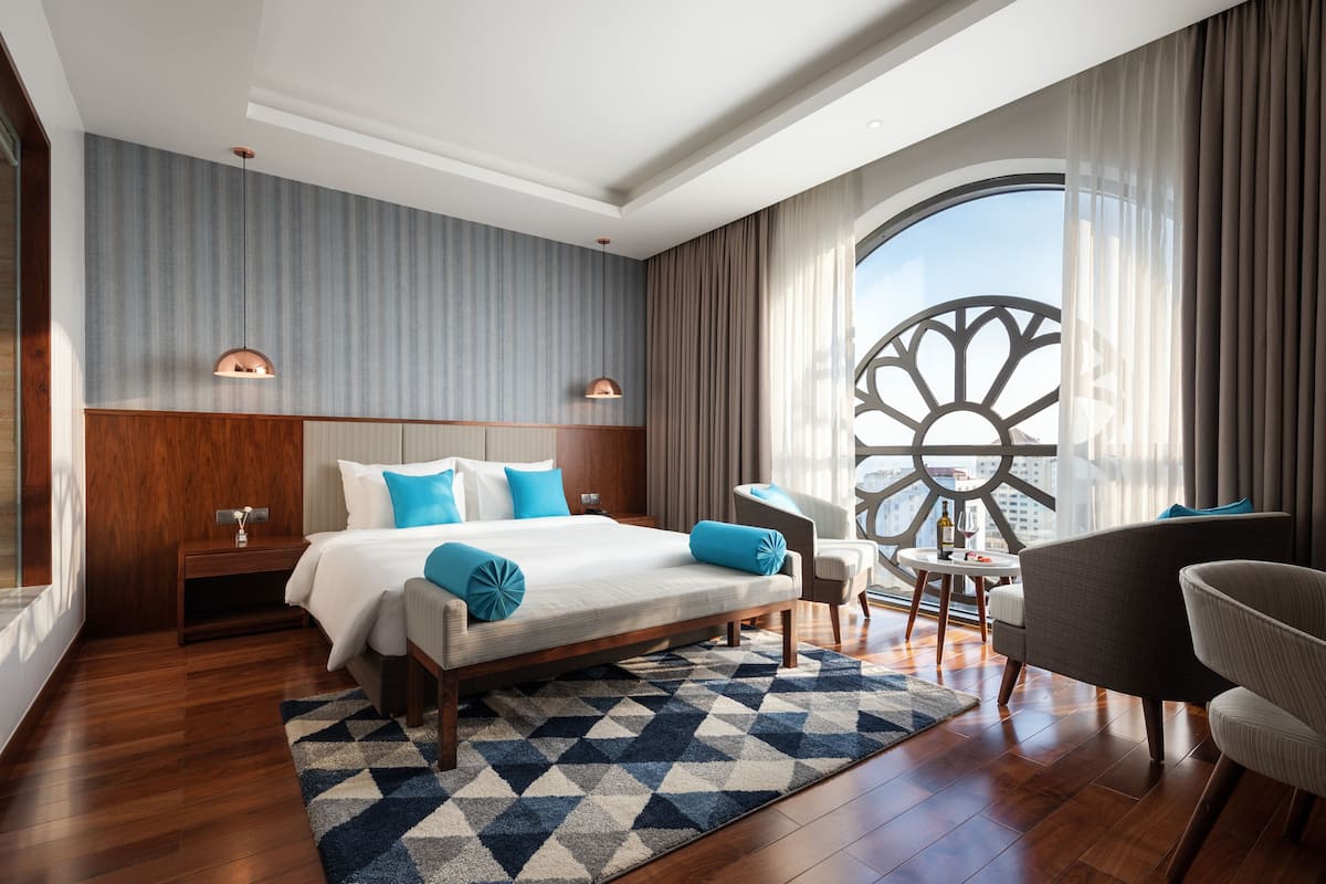 A modern hotel room with a large bed, a seating area with a table, and a view of the city through a large window.