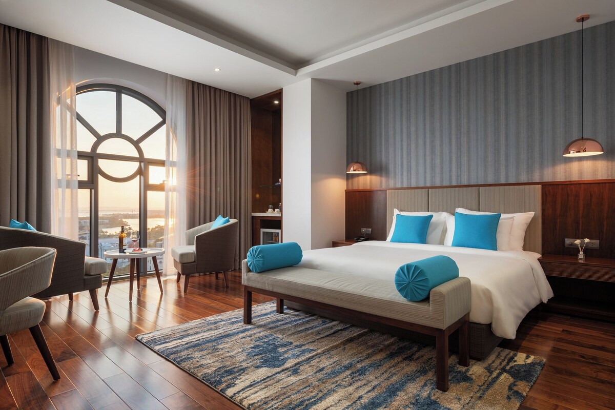 A hotel room with a large bed, a seating area with a table, and a view of the city.