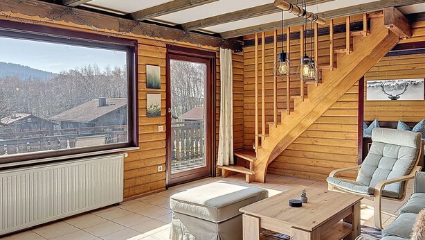 A log cabin interior with a wooden staircase, a large window with a view of a forest, and a cozy seating area with a table and chairs. - Franzi 2 by Interhome (Arrach)