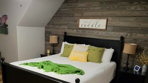 A bedroom with a wooden floor, a bed with a white bedspread, green and yellow pillows, a nightstand with a lamp, and a sign on the wall that says "Let's cuddle".