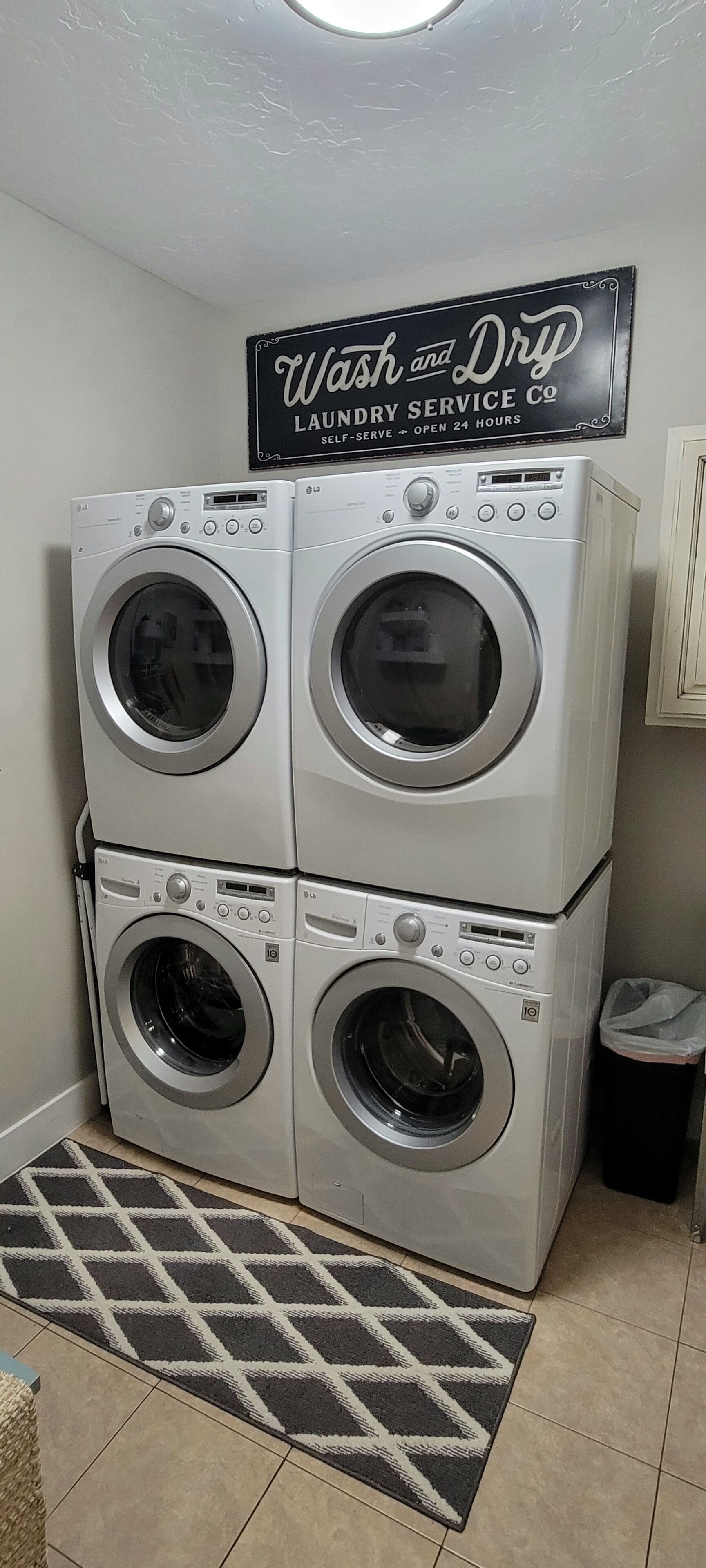 A laundry room with two stacked front-loading washing machines, a sign above them, and a patterned rug on the floor.