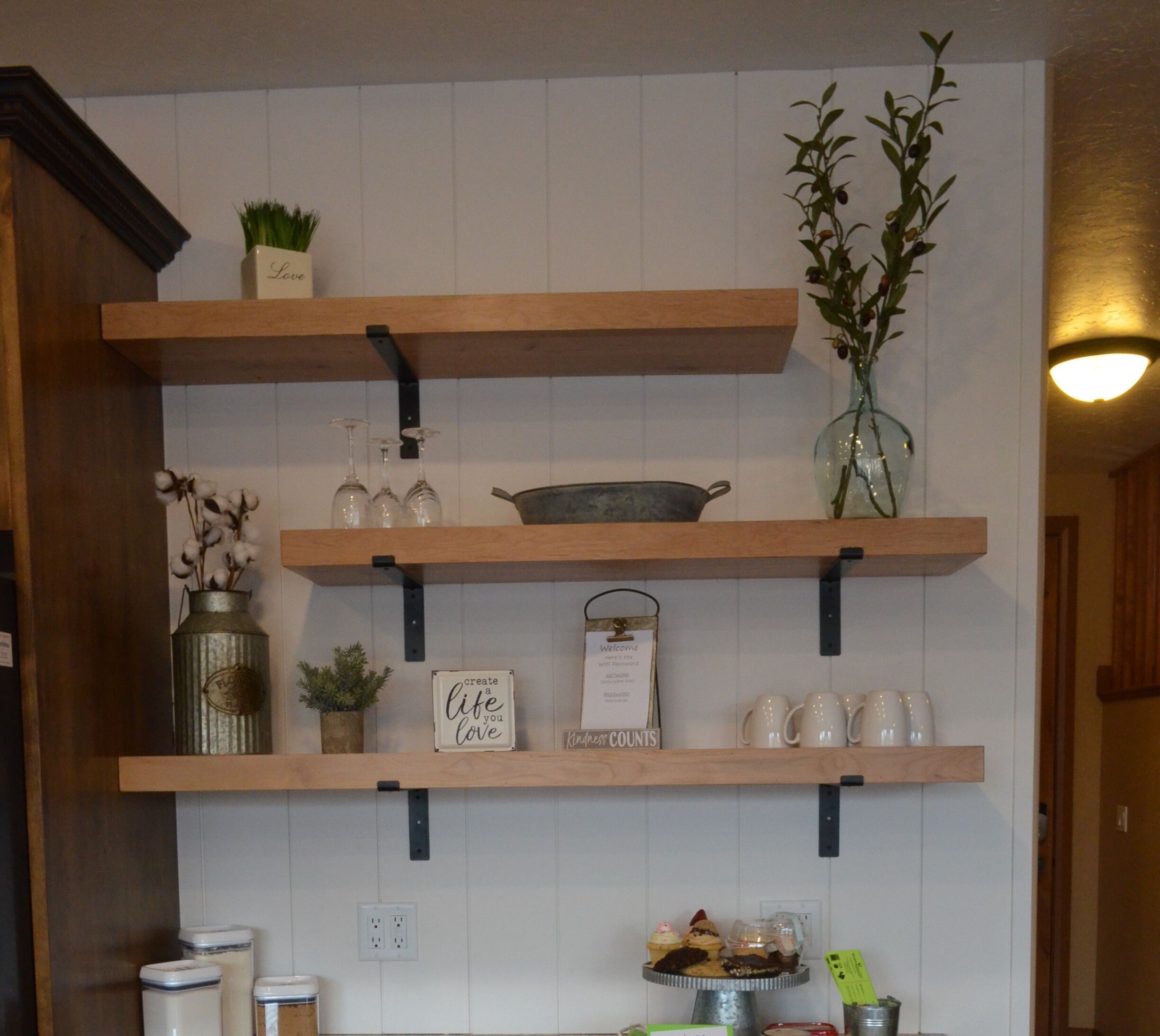 A kitchen with wooden shelves holding various items like a vase, a metal container, and a sign that says "create a life you love."