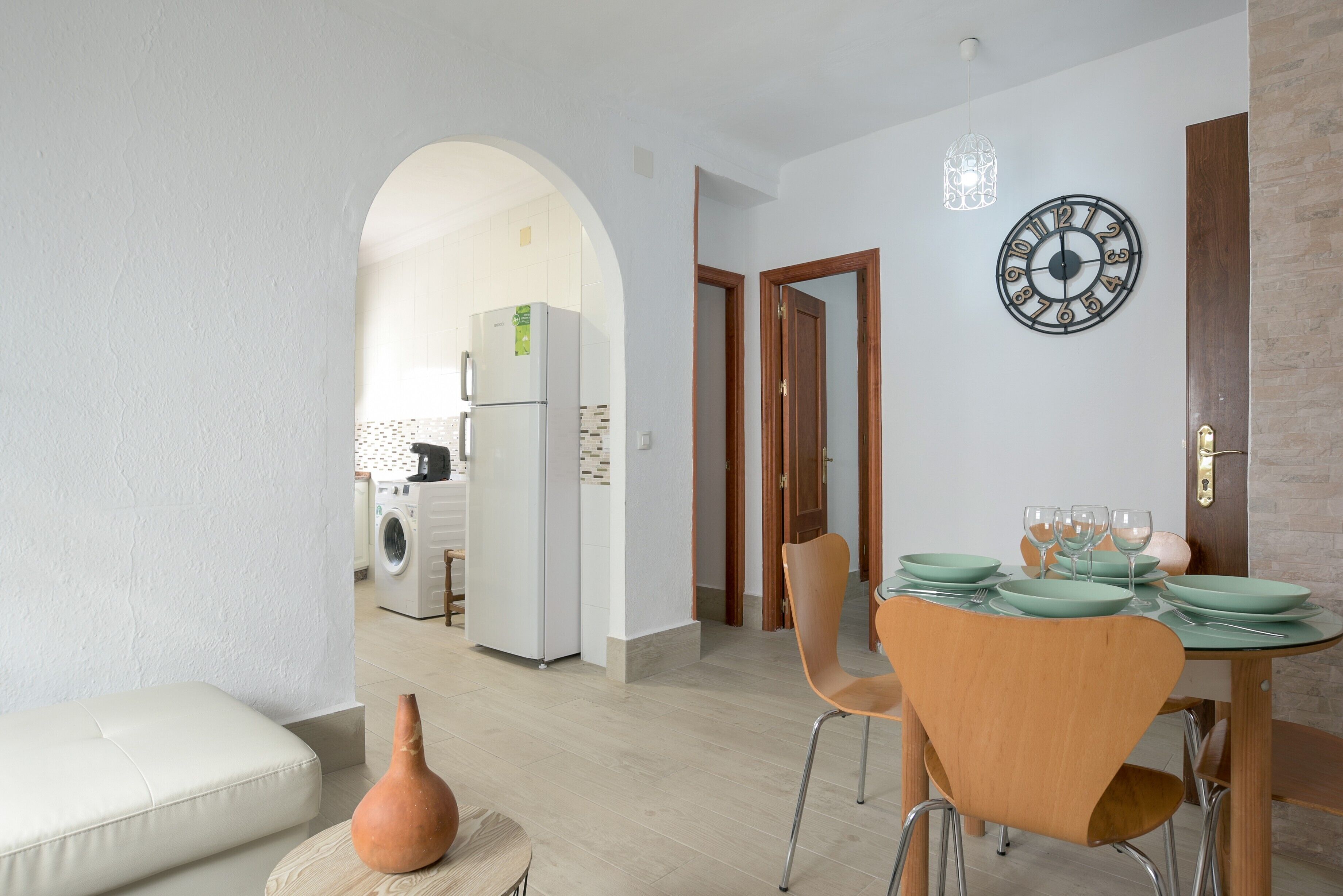 Un comedor con una mesa dispuesta para cuatro personas, un reloj en la pared y vistas a la cocina.