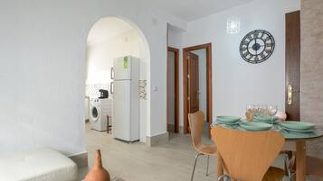 Un comedor con una mesa dispuesta para cuatro personas, un reloj en la pared y vistas a la cocina.
