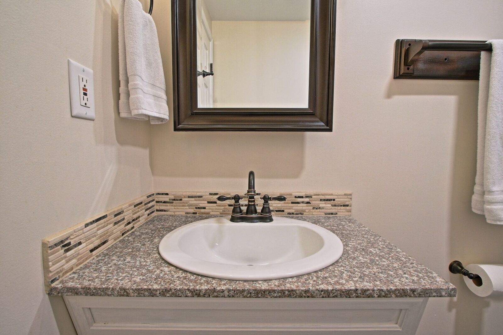A bathroom with a granite countertop sink, a mirror, a towel rack, and a toilet paper holder.