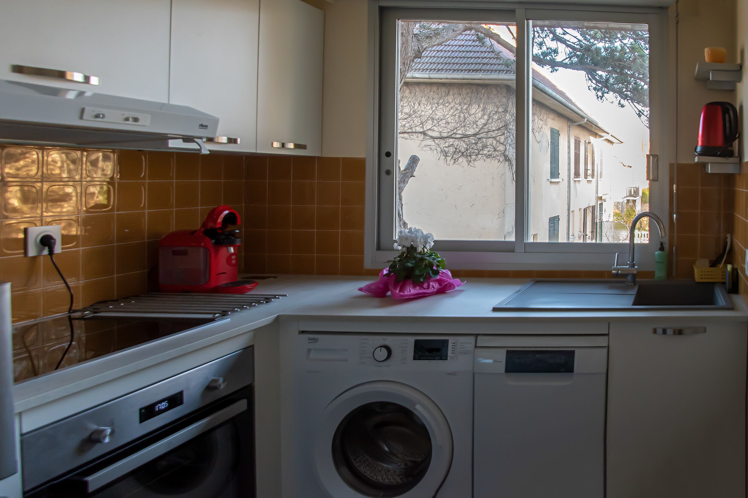 A compact kitchen with a washing machine, a red kettle, and a window with a view of a house.