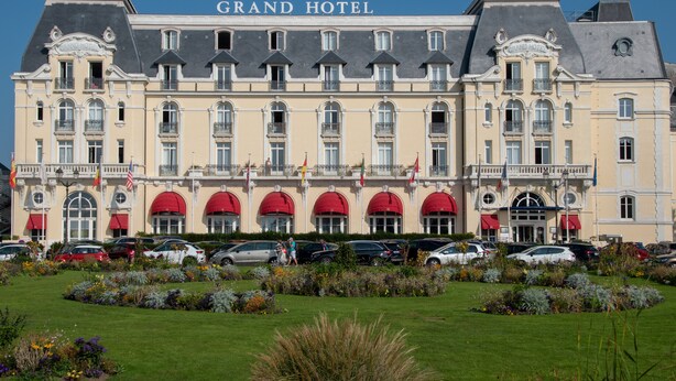 Ein großes, historisches Gebäude mit der Aufschrift "Grand Hotel", einer symmetrischen Fassade, mehreren Balkonen und einem gepflegten Garten davor.
