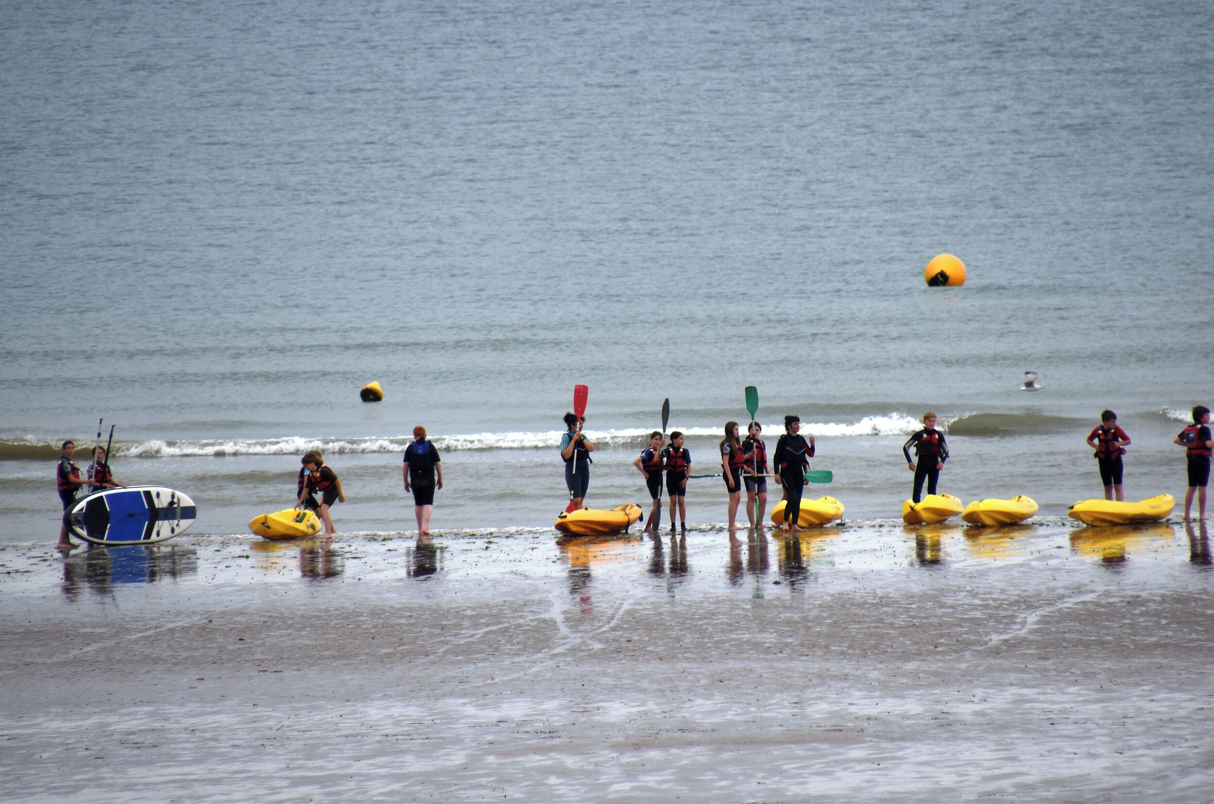 Eine Strandansicht mit Menschen am Ufer, manche davon mit Surfbrettern und Kajaks.