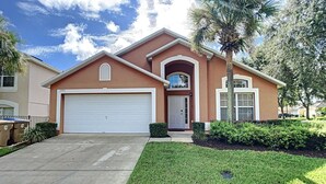A two-story house with a white garage door, a front entrance with a door, and a small front yard with grass and a palm tree. - Emerald Island Resort 4 bed 3 bath with pool/spa (Kissimmee)
