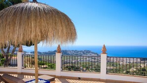 Un balcón con sombrilla de paja, sillones de descanso y vista al mar y al paisaje.
