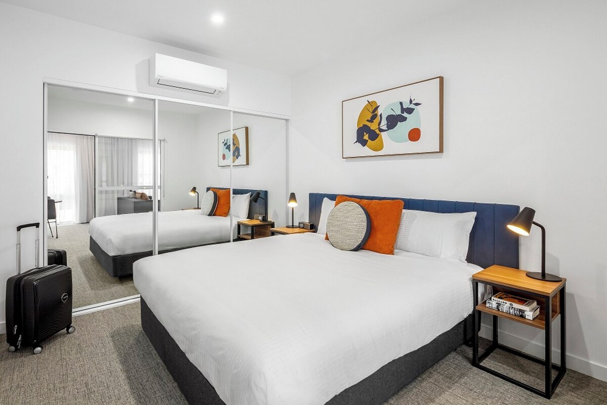 A hotel room with two beds, a large mirror, a bedside table with a lamp, and a wall-mounted artwork.