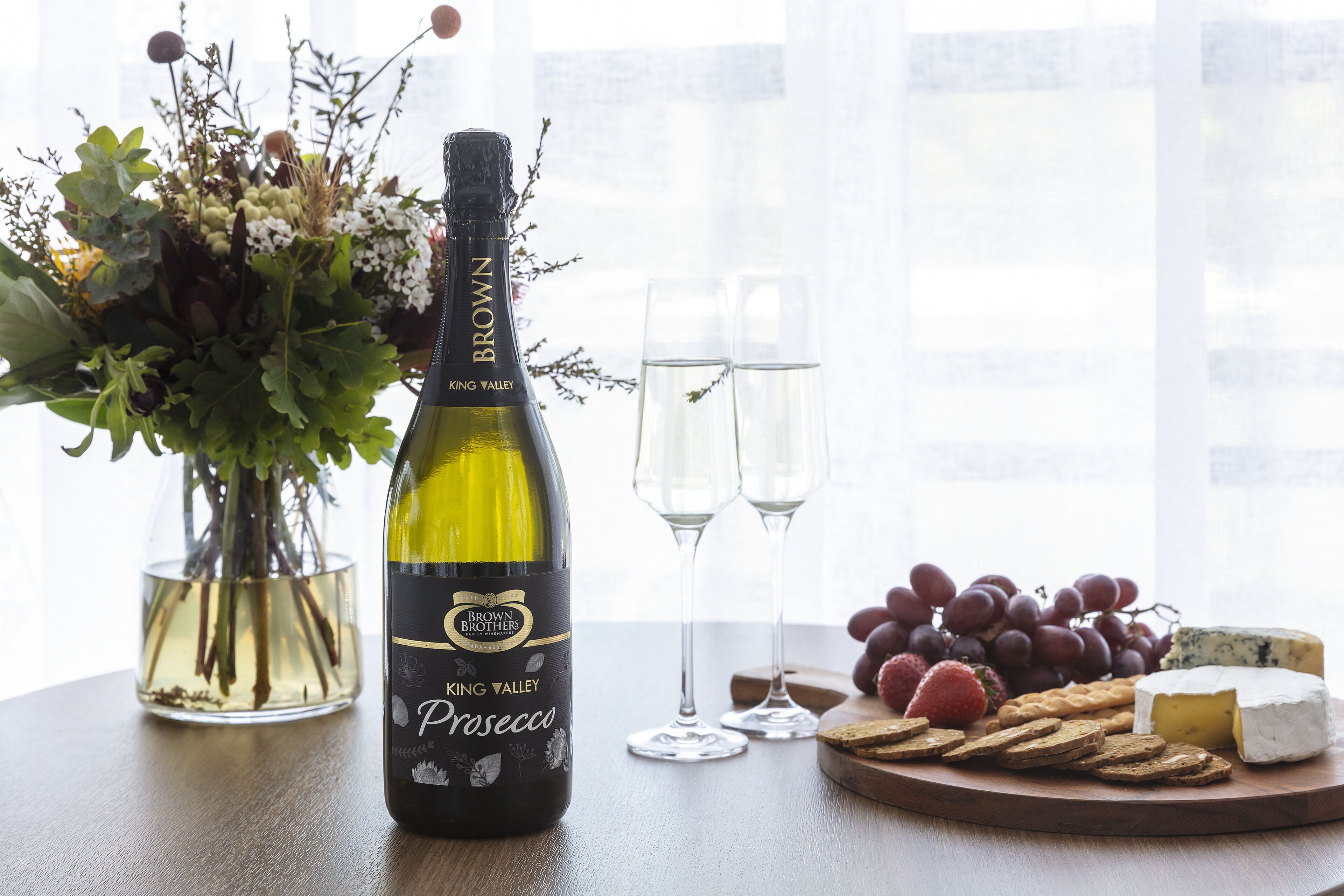 A bottle of Brown Brothers King Valley Prosecco, two glasses, a variety of cheeses, grapes, and crackers on a wooden board.