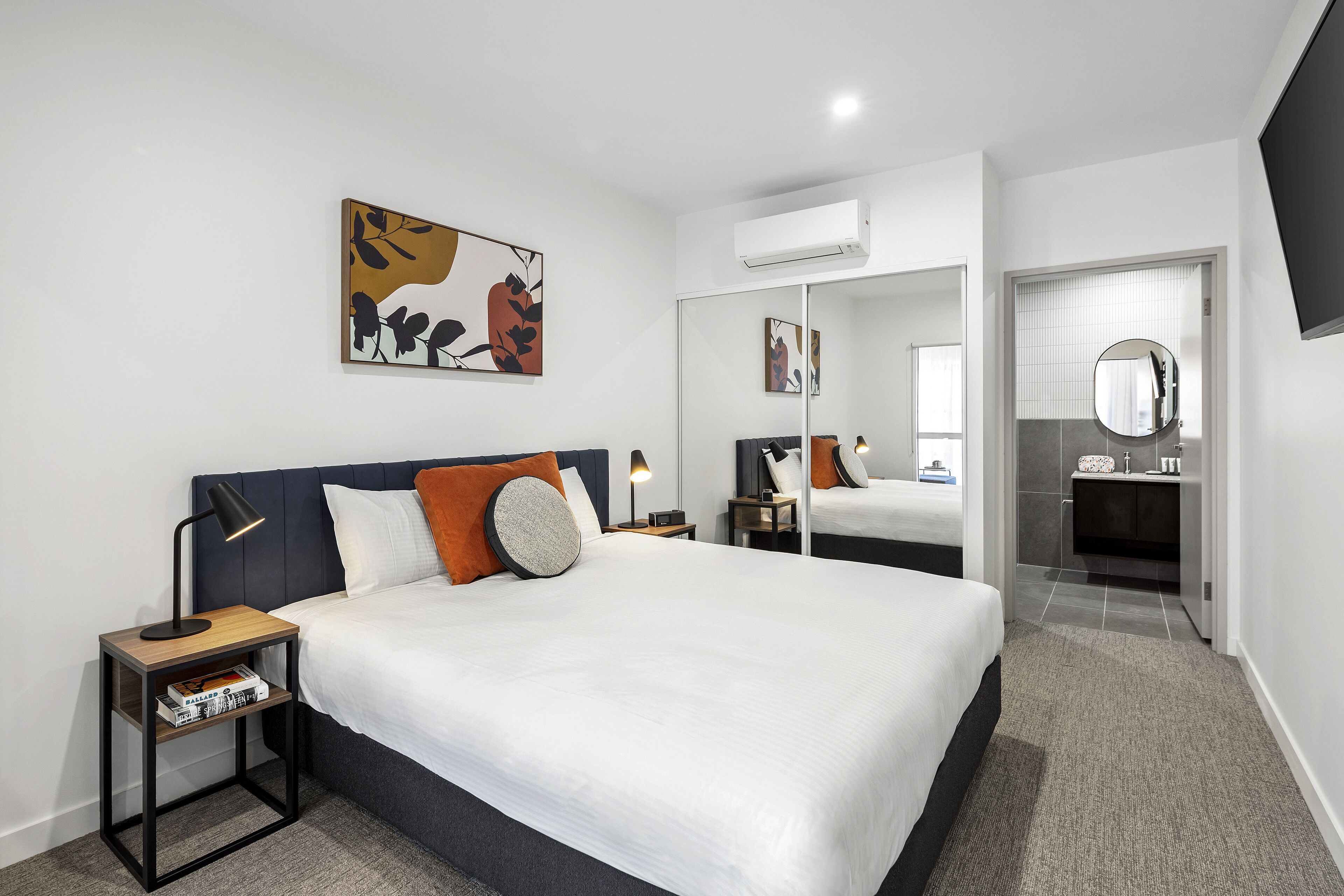 Habitación de hotel moderna con una cama grande, una mesita de noche con lámpara, aire acondicionado de pared, un espejo y un cuadro.