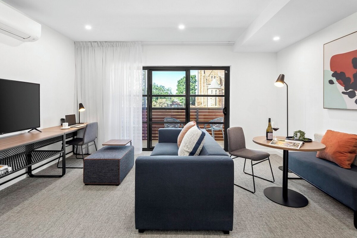 A modern living room with a sofa, a coffee table, a TV, and a large window with a view of a building.