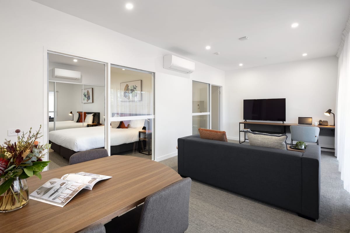 A modern hotel room with a bed, a sofa, a dining table, and a TV.