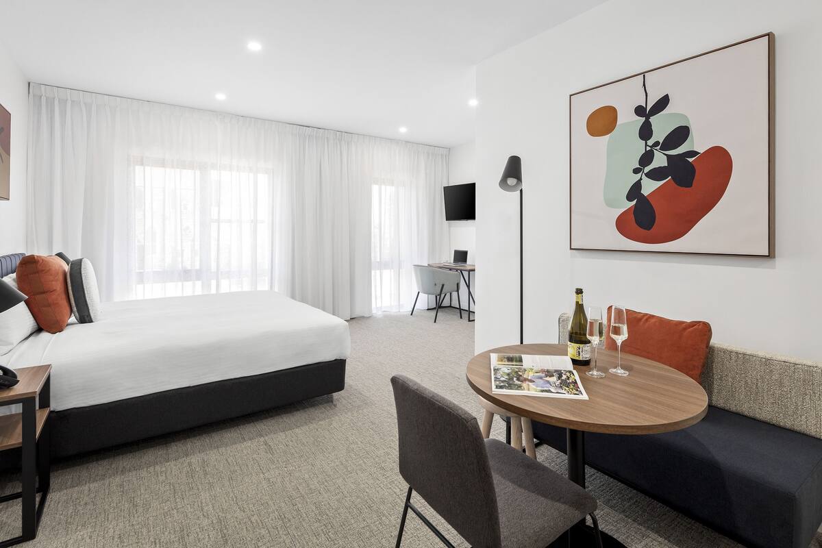 A modern hotel room with a bed, a desk, a round table with a chair, a sofa, and a large abstract painting on the wall.