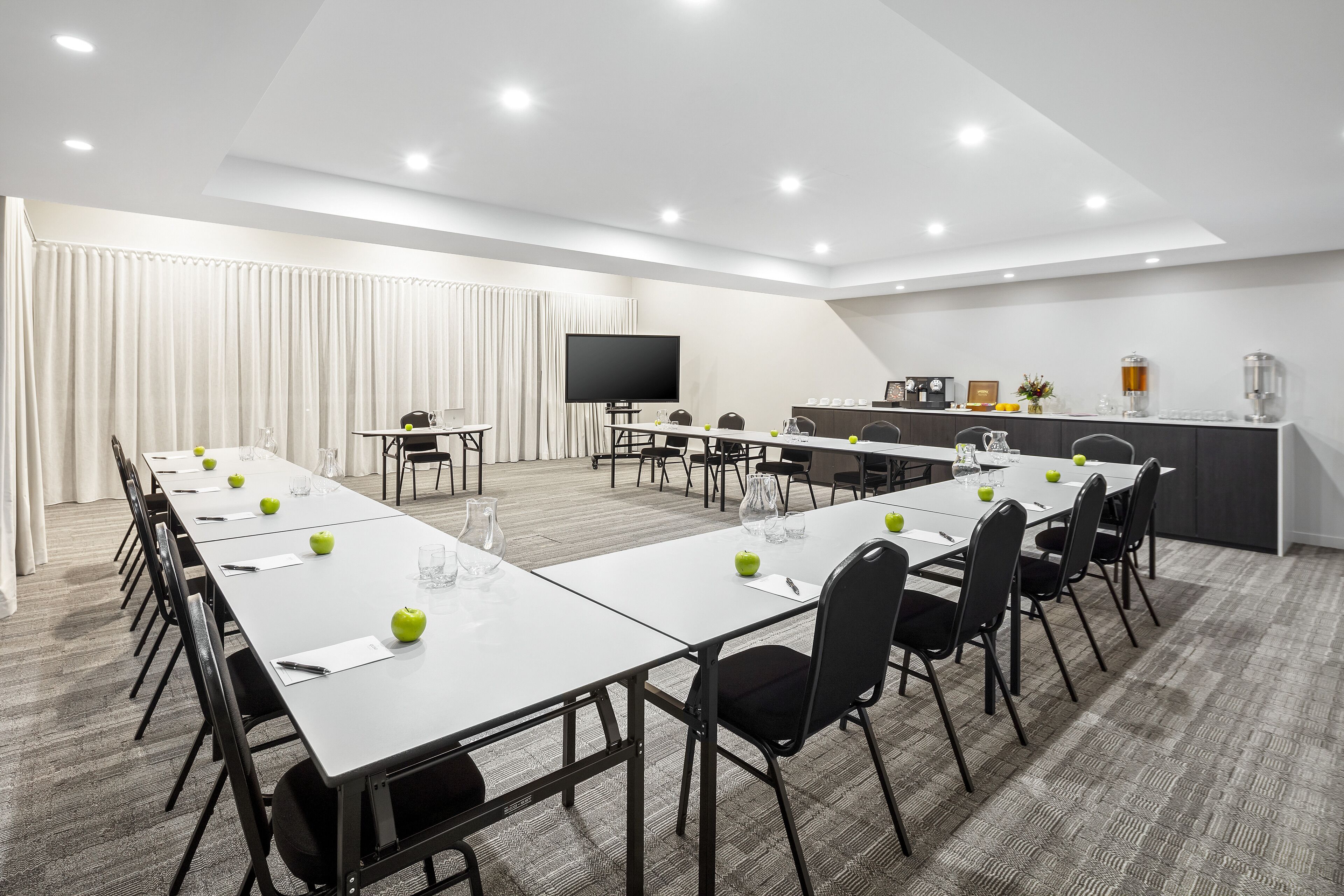 A conference room with a long table, chairs, a TV, and a kitchenette area.