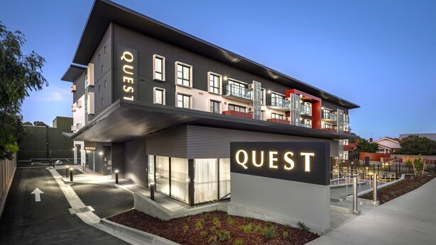 Un edificio moderno con la señalización "QUEST", que presenta una estructura de varios pisos con balcones y ventanas amplias.
