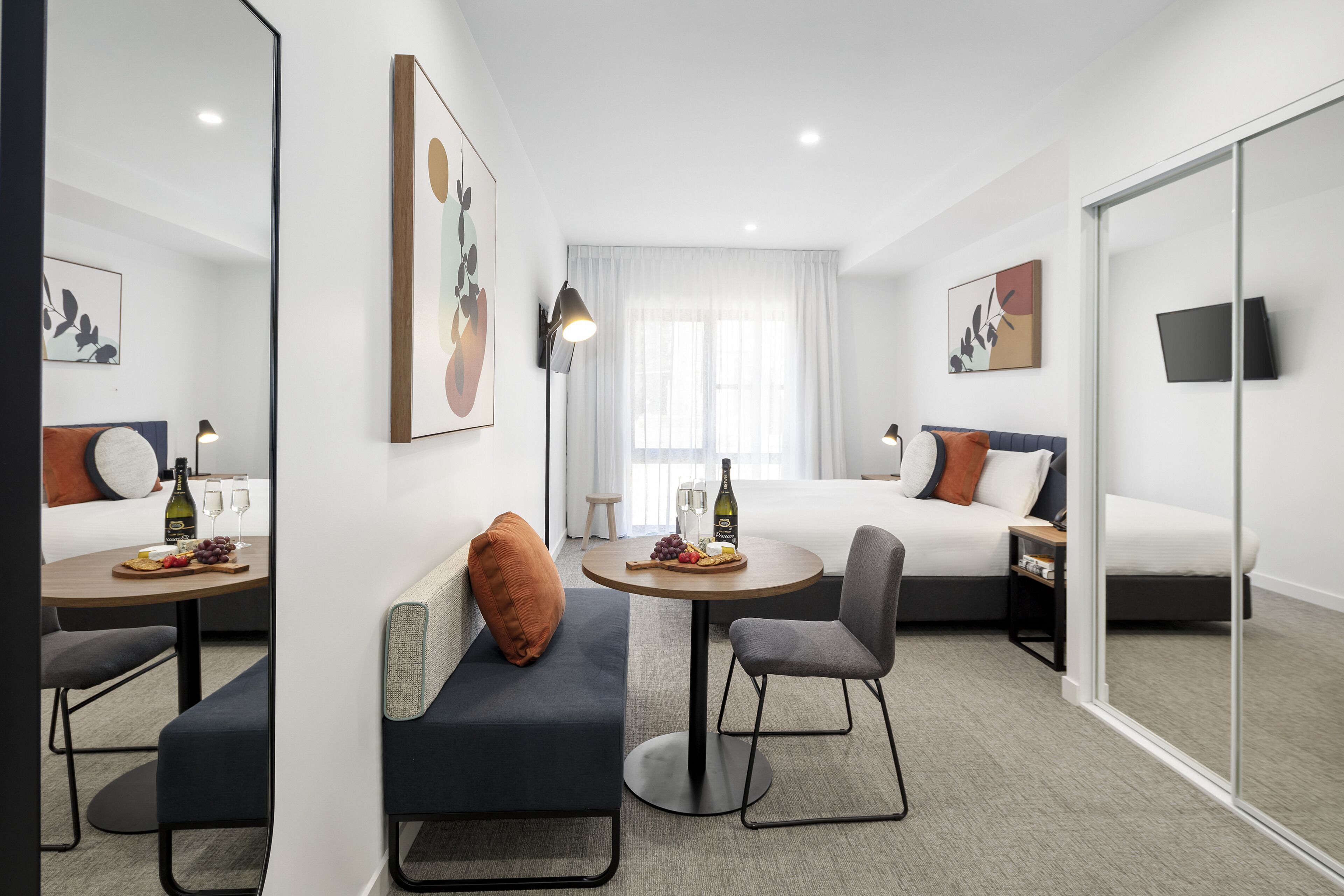 A modern hotel room with a bed, a sofa, a dining table with a bottle and glasses, and a TV on the wall.