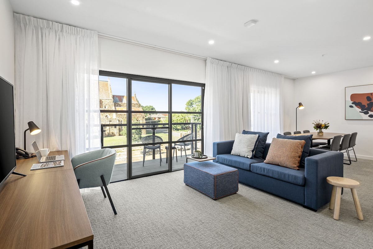 A modern living room with a blue sofa, a wooden desk, and a large sliding glass door leading to an outdoor area.