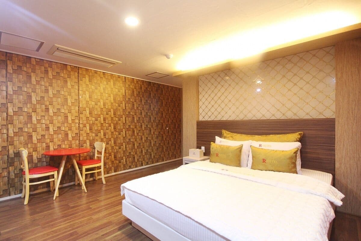 A hotel room with a wooden wall, a bed with white bedding, two red chairs, and a small table.