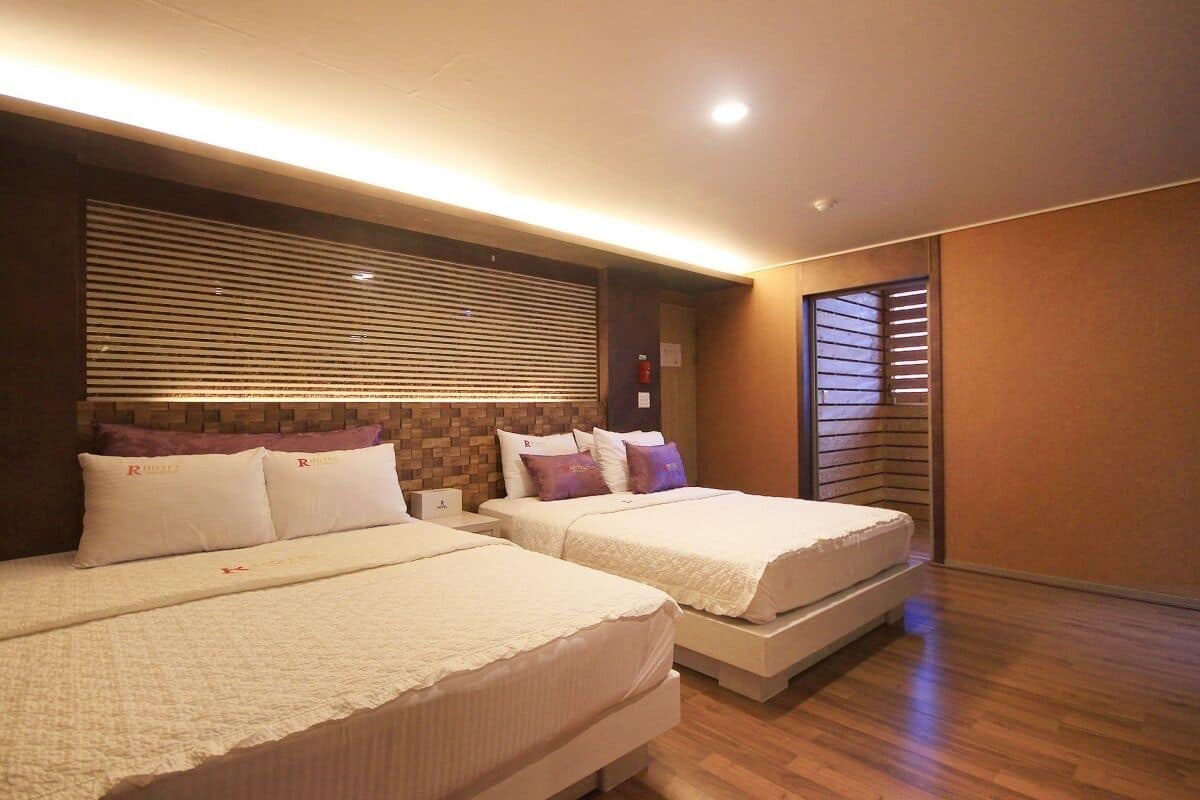 A hotel room with two beds, a brick accent wall, and a window with blinds.