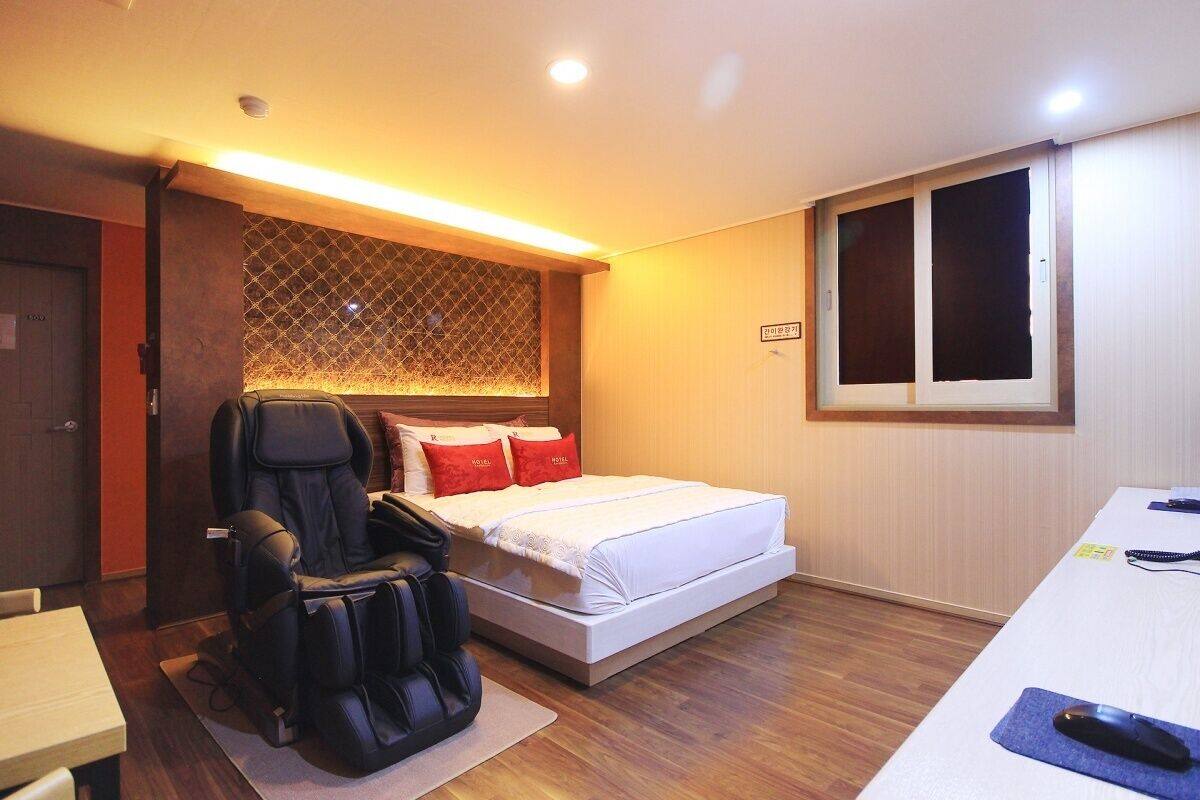 A compact hotel room with a single bed, a massage chair, a desk, and a window with blinds.