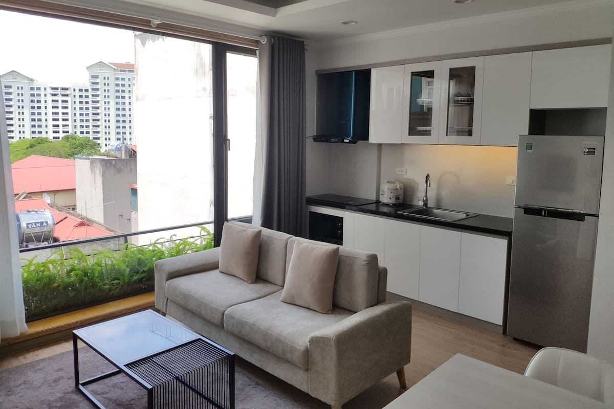 A modern apartment with a sofa, a dining table, a kitchen with white cabinets, and a large window overlooking a cityscape.