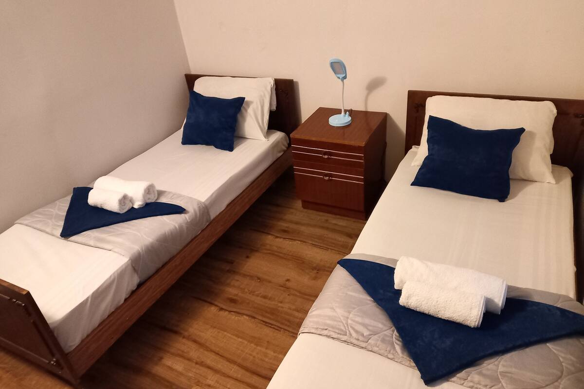 Habitación de hotel con dos camas individuales, cada una con una almohada azul y una manta blanca. Entre las camas hay una mesita de noche de madera con una lámpara.
