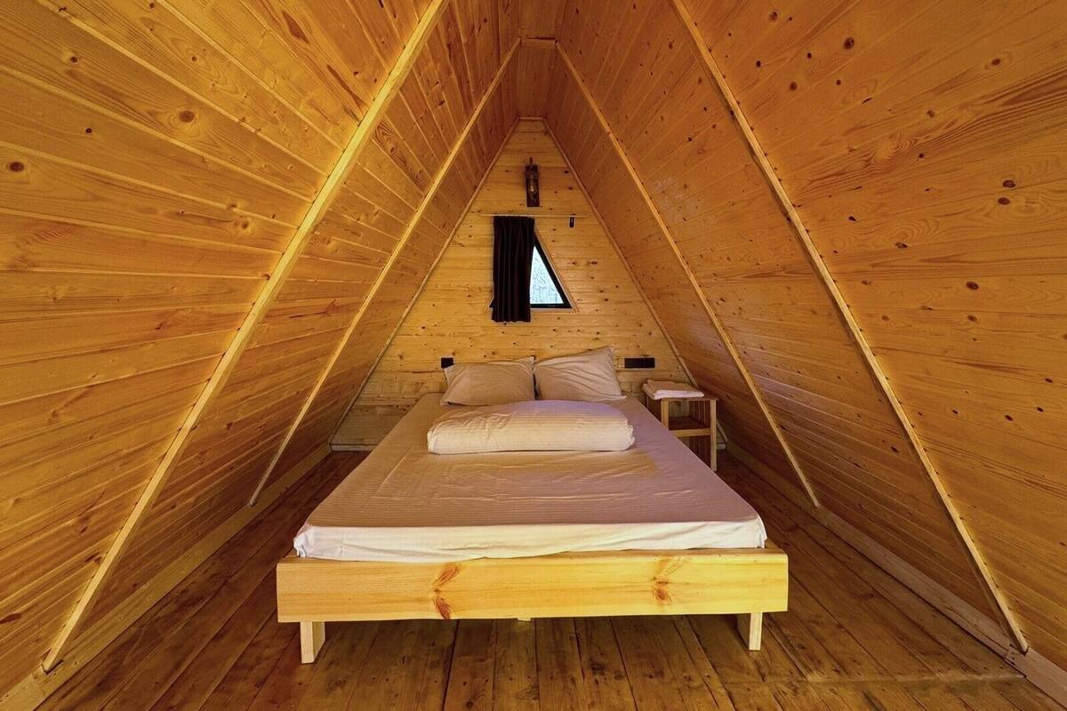 Una habitación de madera con forma triangular, una cama, una mesita y una ventana.