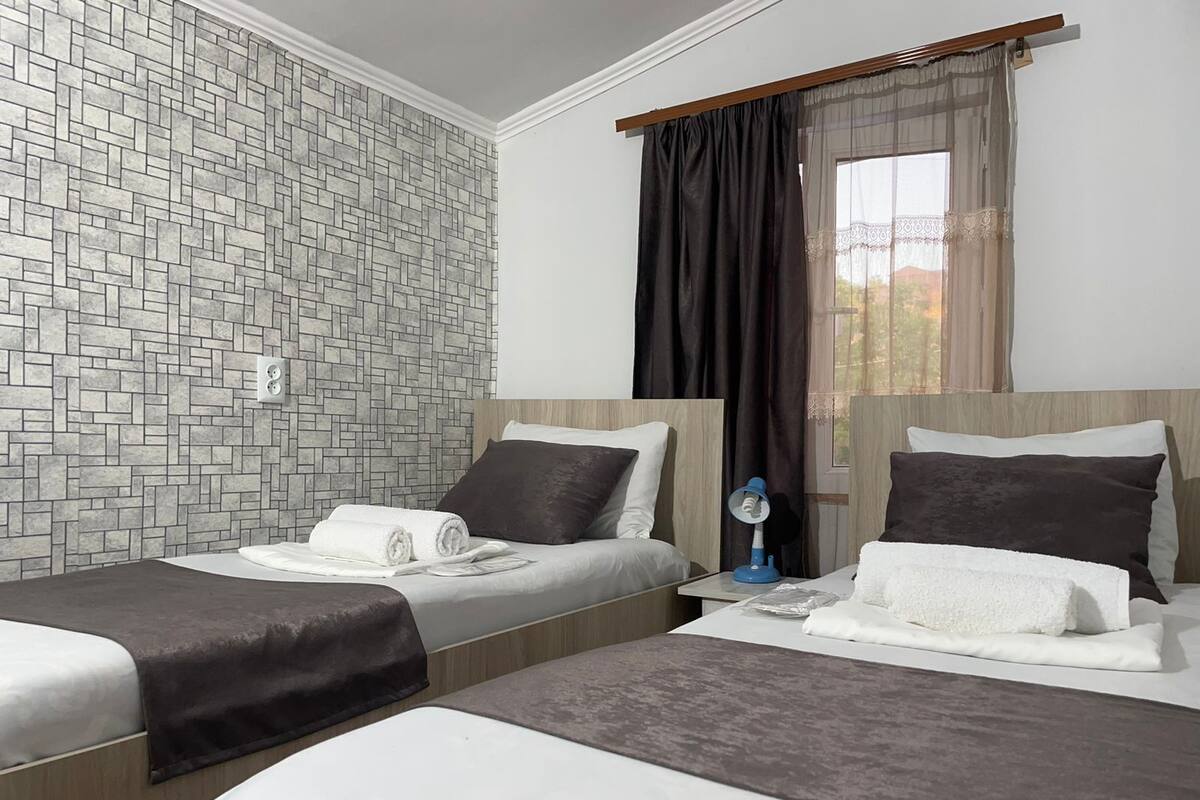 Habitación de hotel con dos camas, una pared con diseño de ladrillos, una ventana con cortinas translúcidas y una mesita de noche con lámpara.