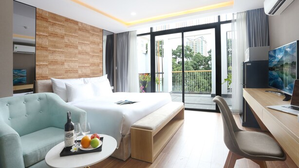 Une chambre d’hôtel moderne équipée d’un lit, d’un canapé, d’un bureau et d’une télévision. Elle dispose également d’un balcon offrant une vue sur les bâtiments et les arbres.
