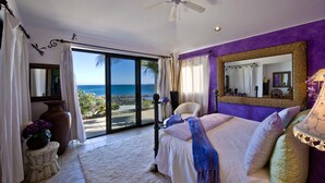 Quarto com cama, ventoinha no teto, janela grande com vista para o oceano e parede roxa.