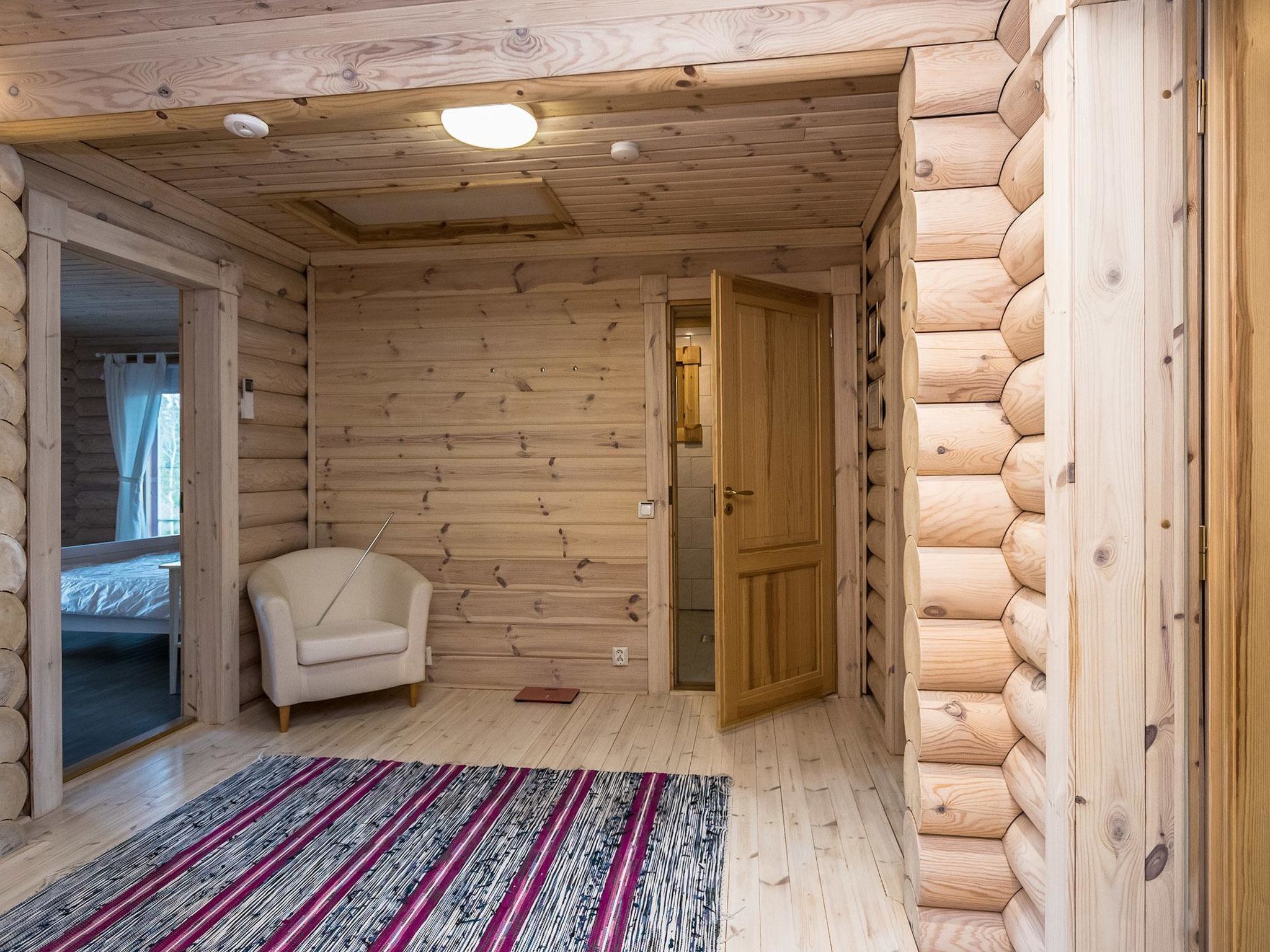 A log cabin room with a bed, a chair, a wooden floor, and a door leading to another room.